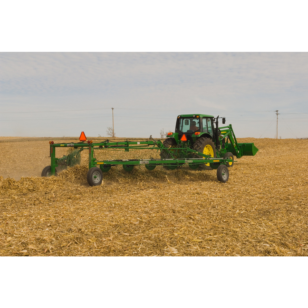 Rear right view of a tractor pulling a WR1212 High-Capacity Wheel Rake through a tan, harvested field.