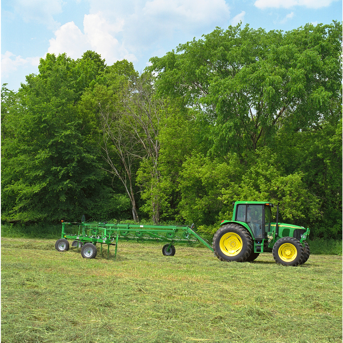 Side right view of a tractor using a green WR High-Capacity Wheel Rake in a hay field bordered by trees