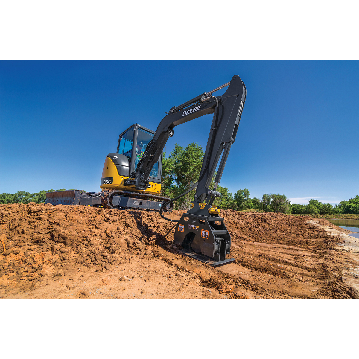 Yellow and black tracked excavator using a plate compactor attachment to smooth dirt on a large construction site, with green trees under a clear blue sky in the background