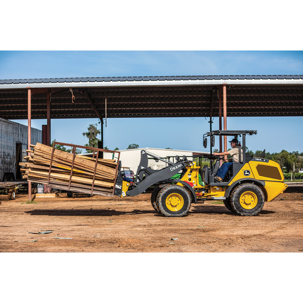 A wheel loader using pallet forks to carry a stacked bundle of lumber across a dirt yard, with an open-sided metal structure and additional materials visible in the background.