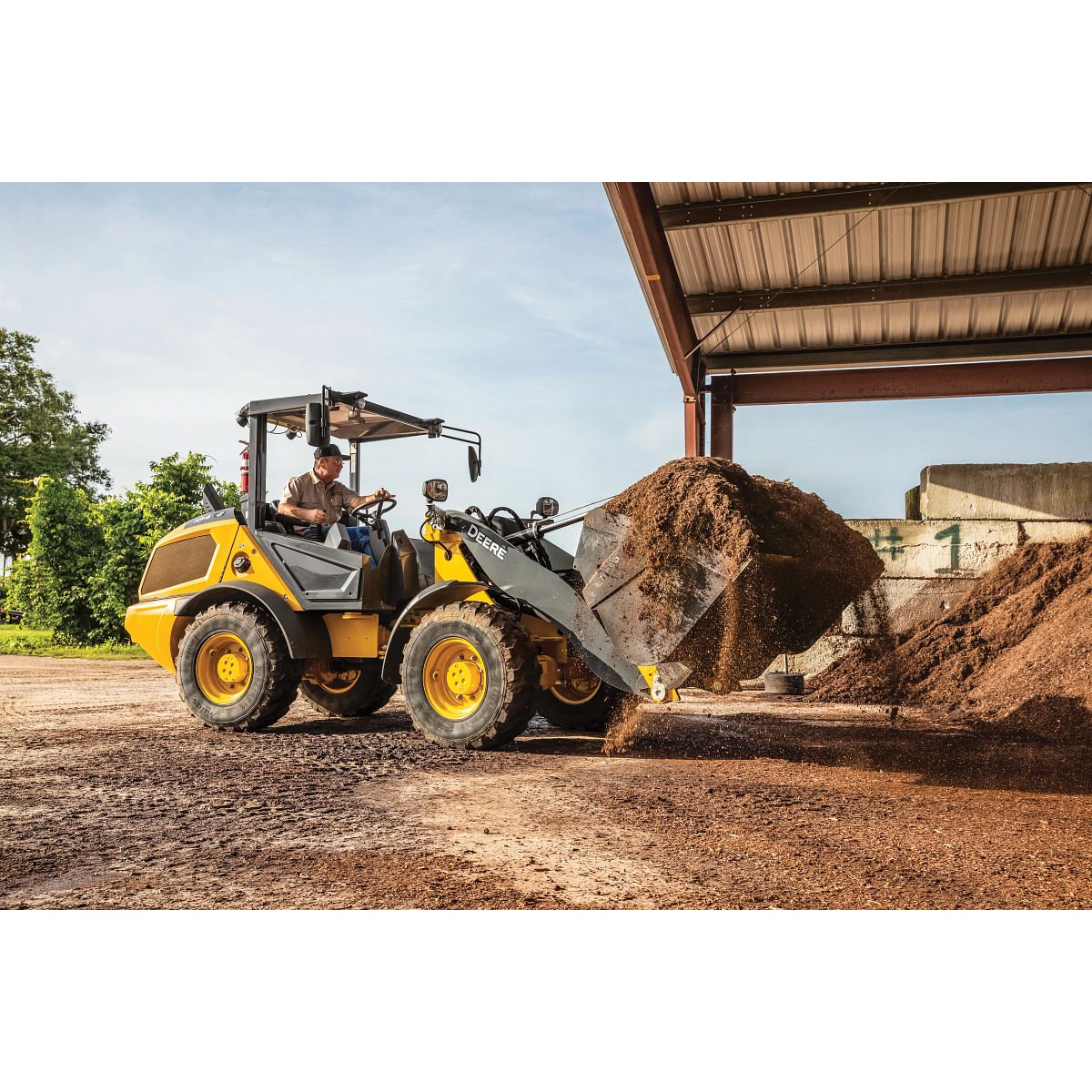 A wheel loader lifting a bucket of loose soil inside an open-sided structure, with material piles on the ground and daylight visible beyond the roofed area.