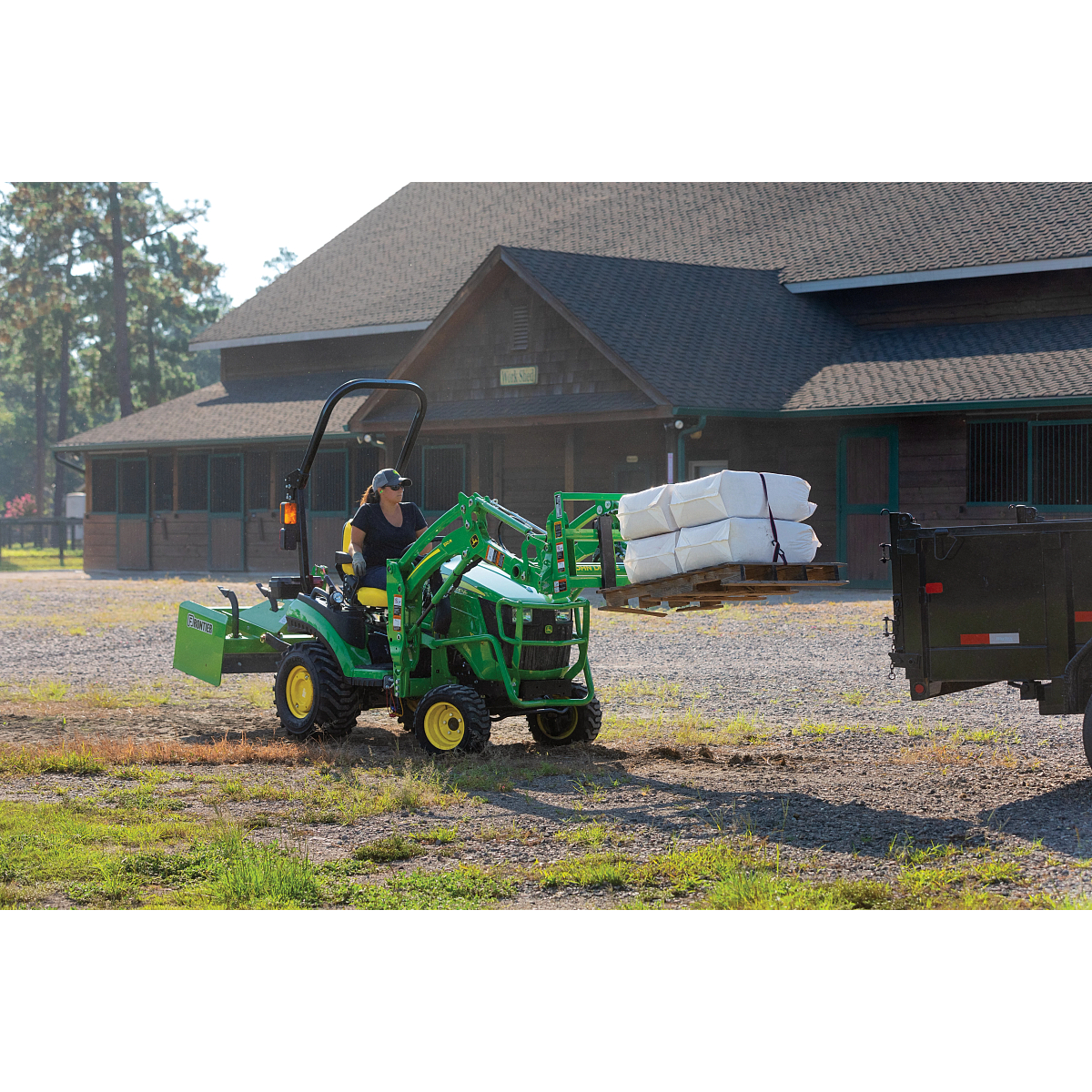 A person operates a small green tractor carrying stacked white bags on the front loader while driving across a gravel area in front of a large horse stable, with a black trailer nearby on a sunny day