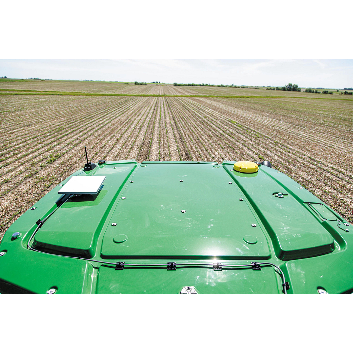 Wide view of a John Deere machine roof showing a mounted JDLink Boost Mini Terminal, antenna, and field of planted crop rows extending to the horizon.