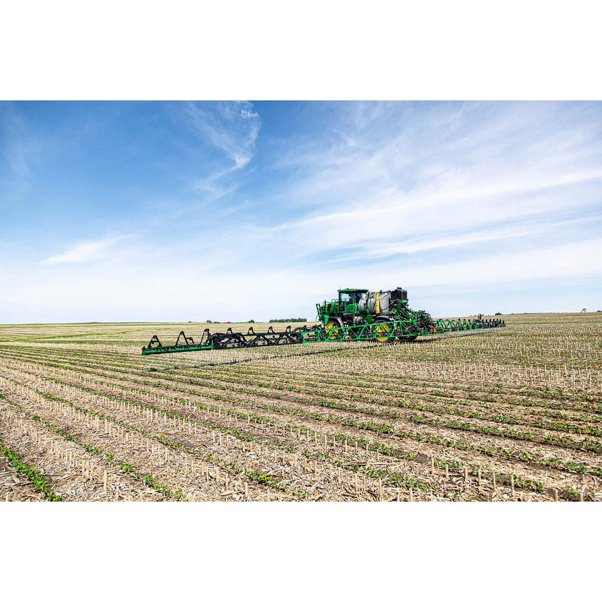 John Deere agricultural machine equipped with a JDLink Boost Mini Terminal operating across a field of crops, beneath a blue sky with white clouds.