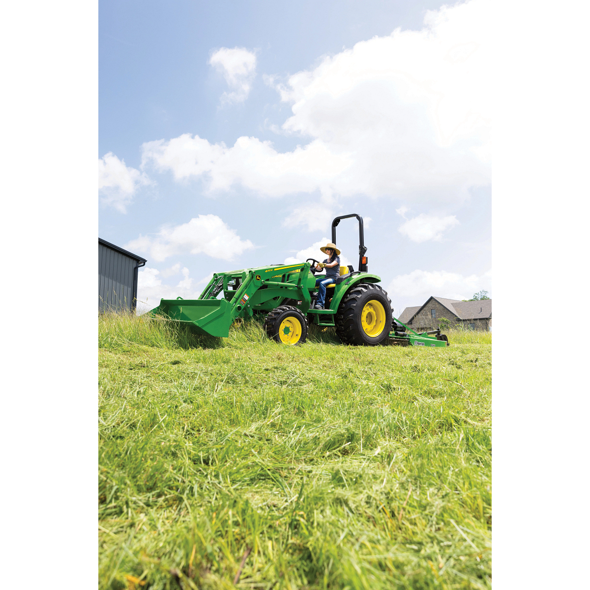 Side left view of a farmer driving a 4044M open operator station tractor across a grassy field. A 400E Loader and RC2072 Rotary Cutter are attached.