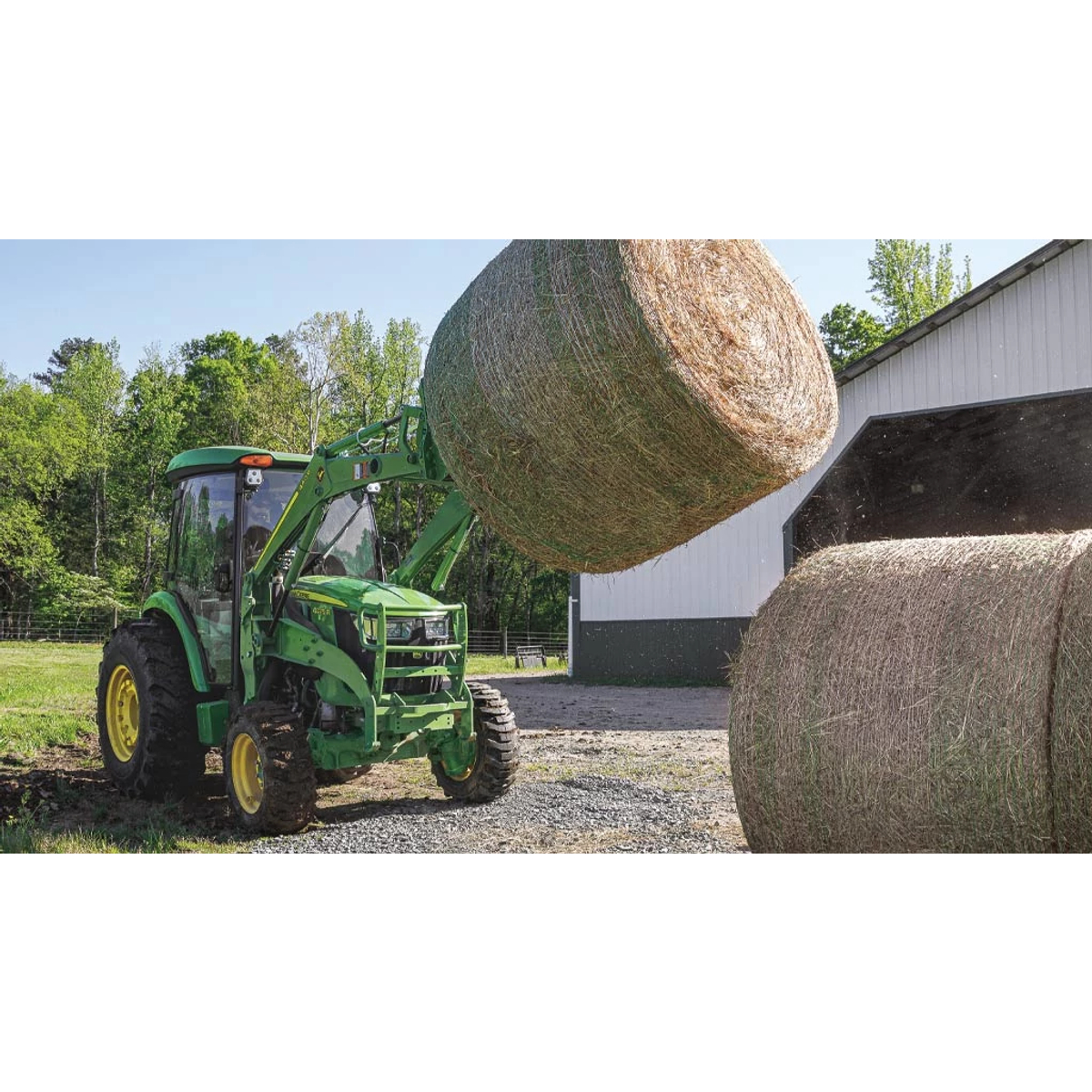 Front right view of a 4R series tractor with front loader lifting a large round hay bale next to another in front of a barn