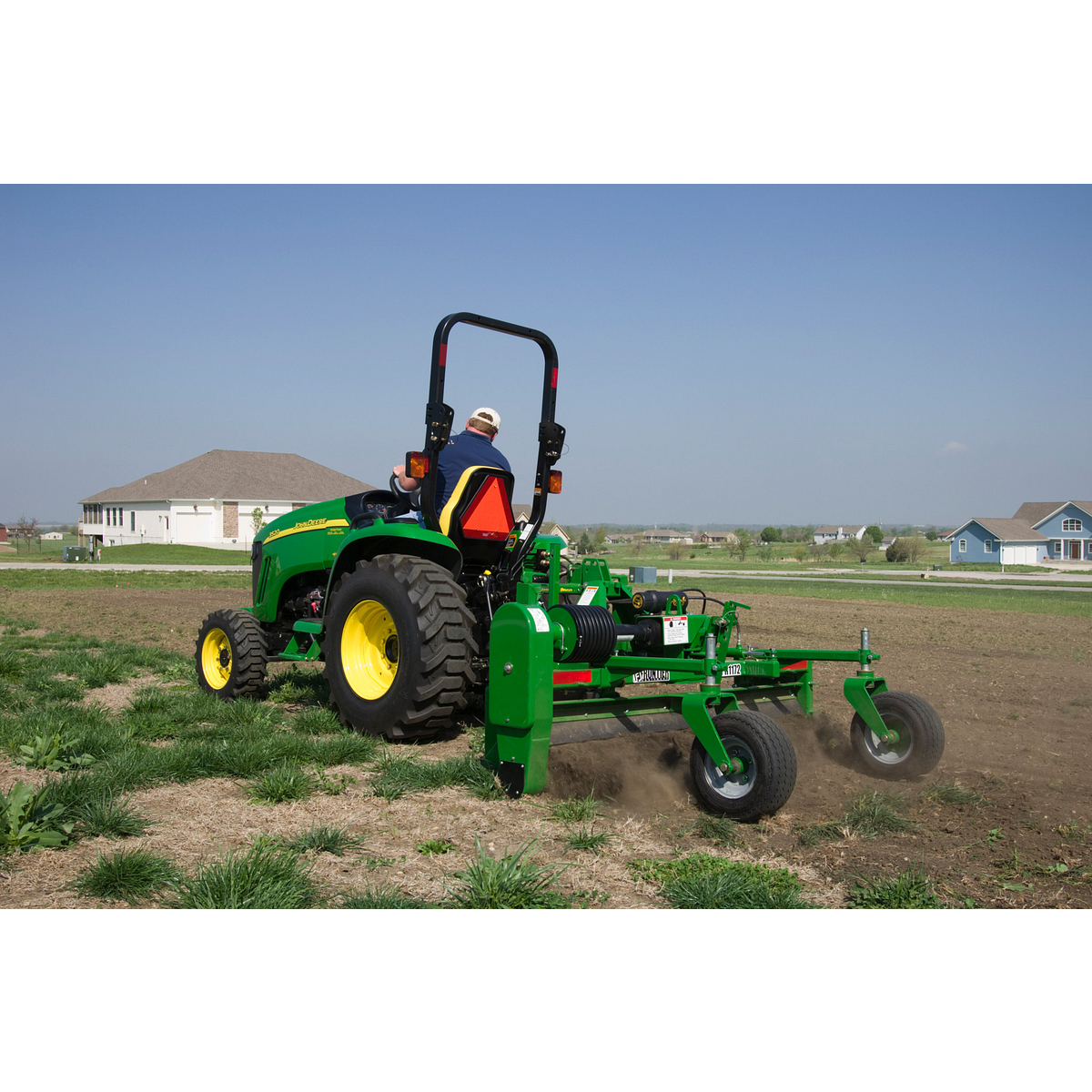 A person operating a green tractor with a rear-mounted soil cultivation attachment, moving across a dry, grassy field. Dirt is being turned behind the implement, with houses and open land visible in the background under a clear sky.