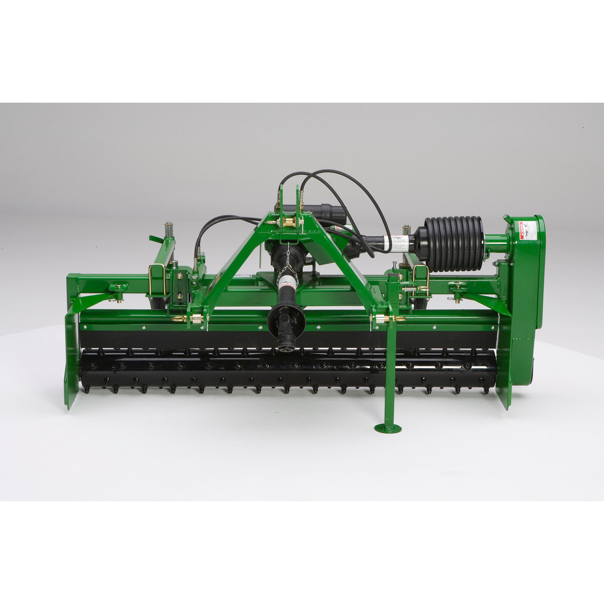 Front view of a rotary tiller or flail mower with PTO shaft, hydraulic hoses, and a wide drum with black blades, shown against a plain light background.