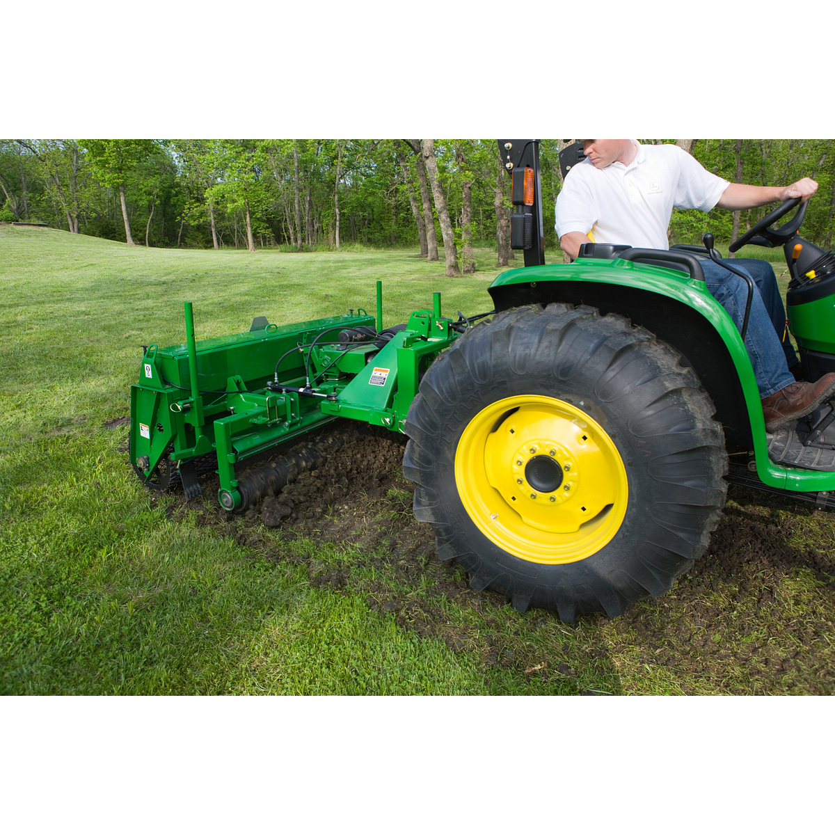 A green tractor with yellow wheels pulls a green soil-cutting attachment that is digging a narrow trench in a grassy field, while a person operates the tractor. Trees and open lawn are visible in the background.