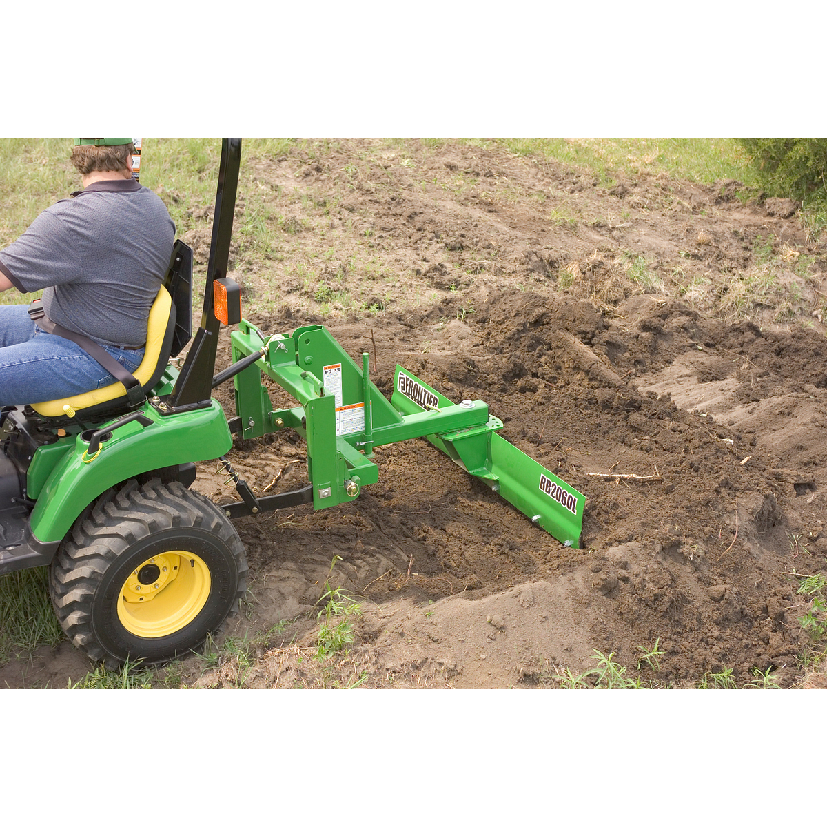 Side view of a green RB2060L Rear Blade at work in dirt, with half of the attached tractor visible and operator atop it turning back to look at the blade.
