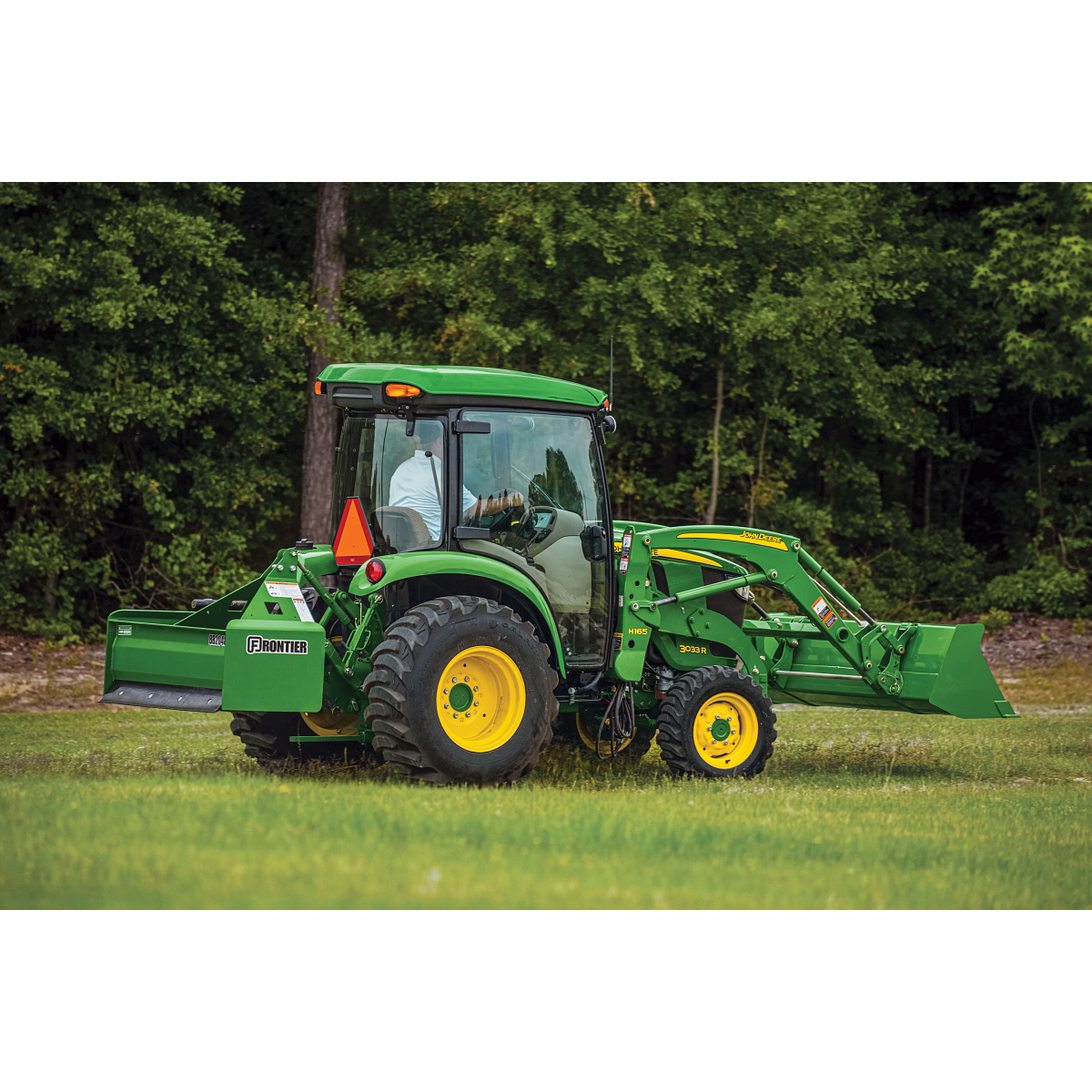 Side view of a green tractor with a front loader driving across a grassy field with an operator inside the enclosed cab and trees forming a dense wooded background