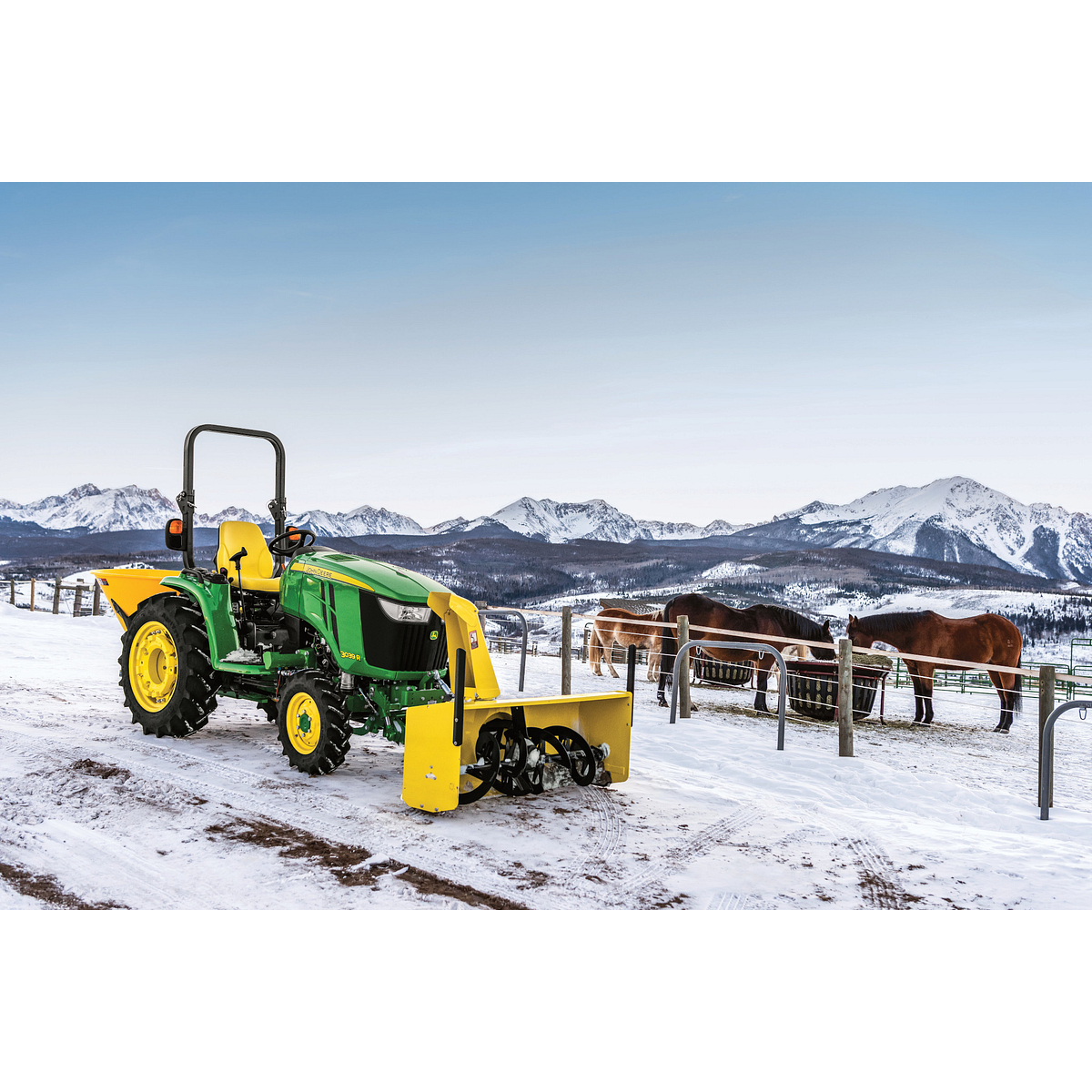 Front right view of an OOS 3039R compact utility tractor parked on a snowy road with horses and mountains in the background and yellow front-attached 60 inch snow blower