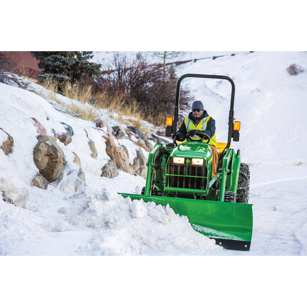 A small green tractor with a front-mounted plow clears a snow-covered road, driven by an operator wearing a high-visibility jacket, with rocks and grass lining the snowy roadside