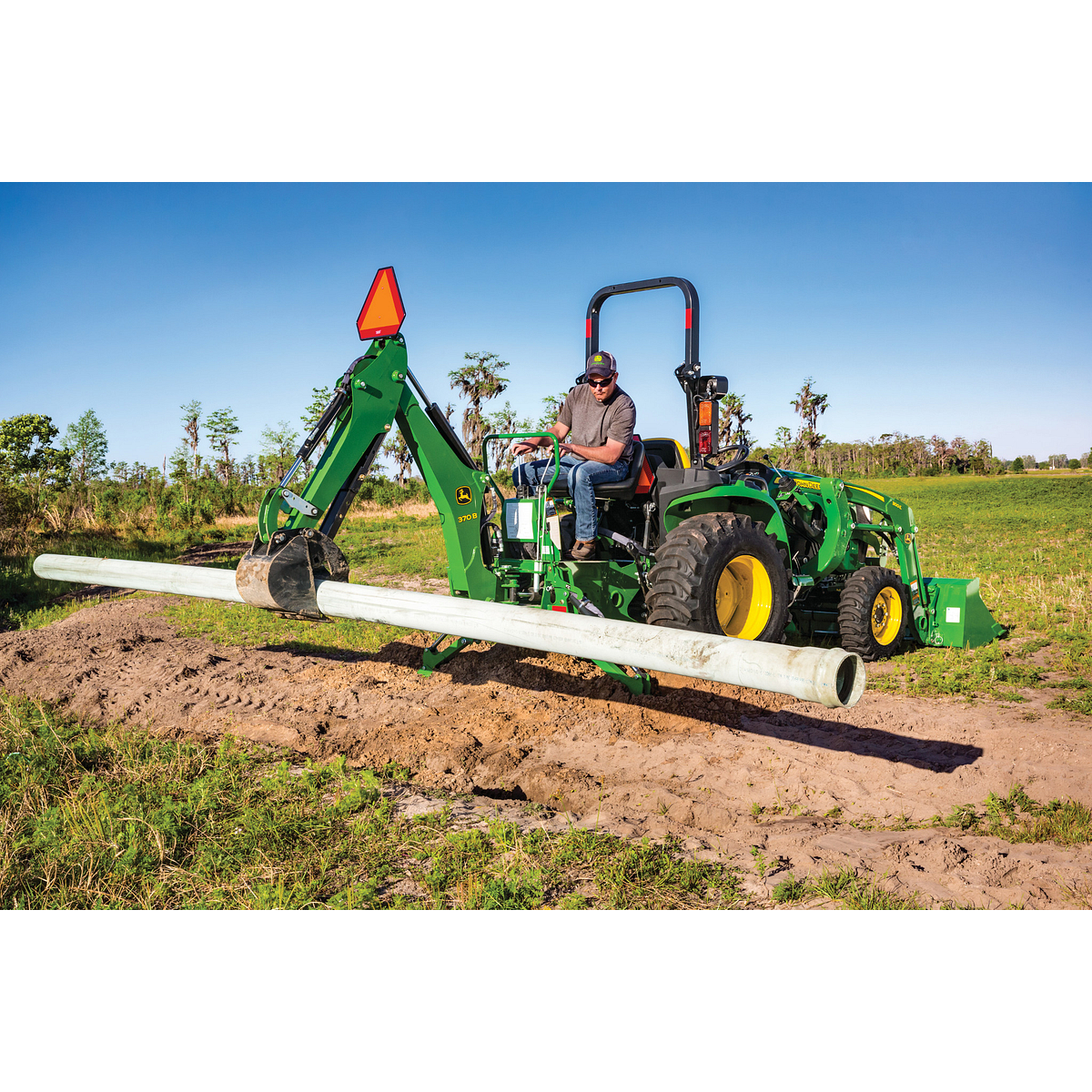 Person operating a green tractor with a backhoe attachment carrying a long pipe over a trench in rural field, with grassy landscape and trees under a clear blue sky in the background