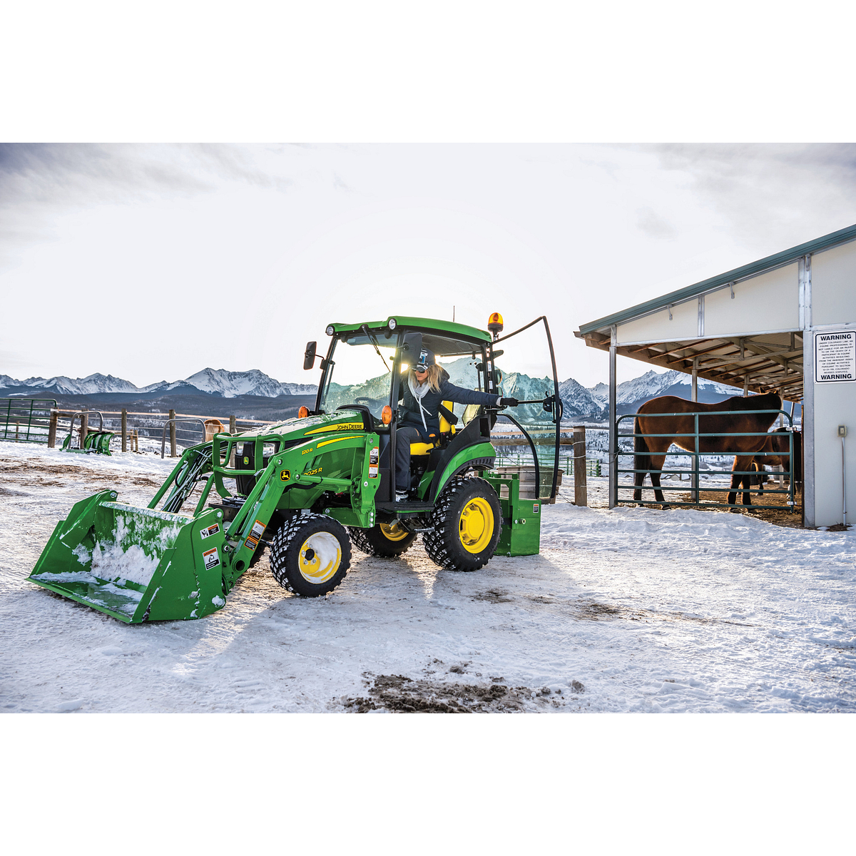 Green tractor with a front loader sitting on a snowy farmyard, as an operator steps out near a barn and fenced livestock area under a cloudy winter sky