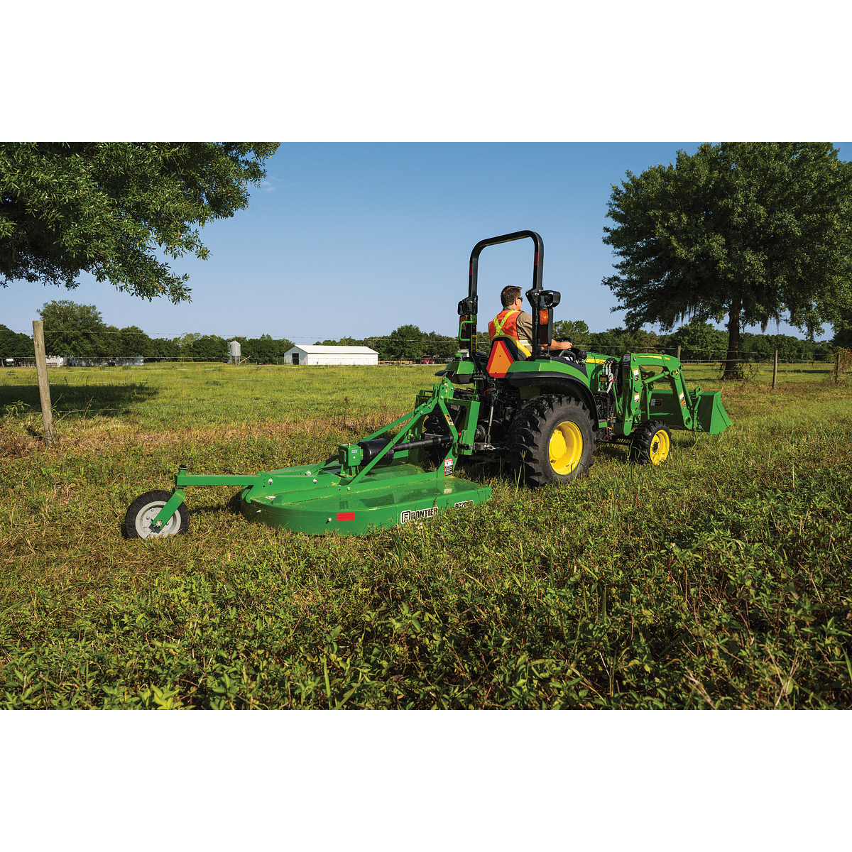 Person operating a green tractor pulling a wide mower across a grassy field, cutting vegetation with trees and a white barn in the background under a clear blue sky