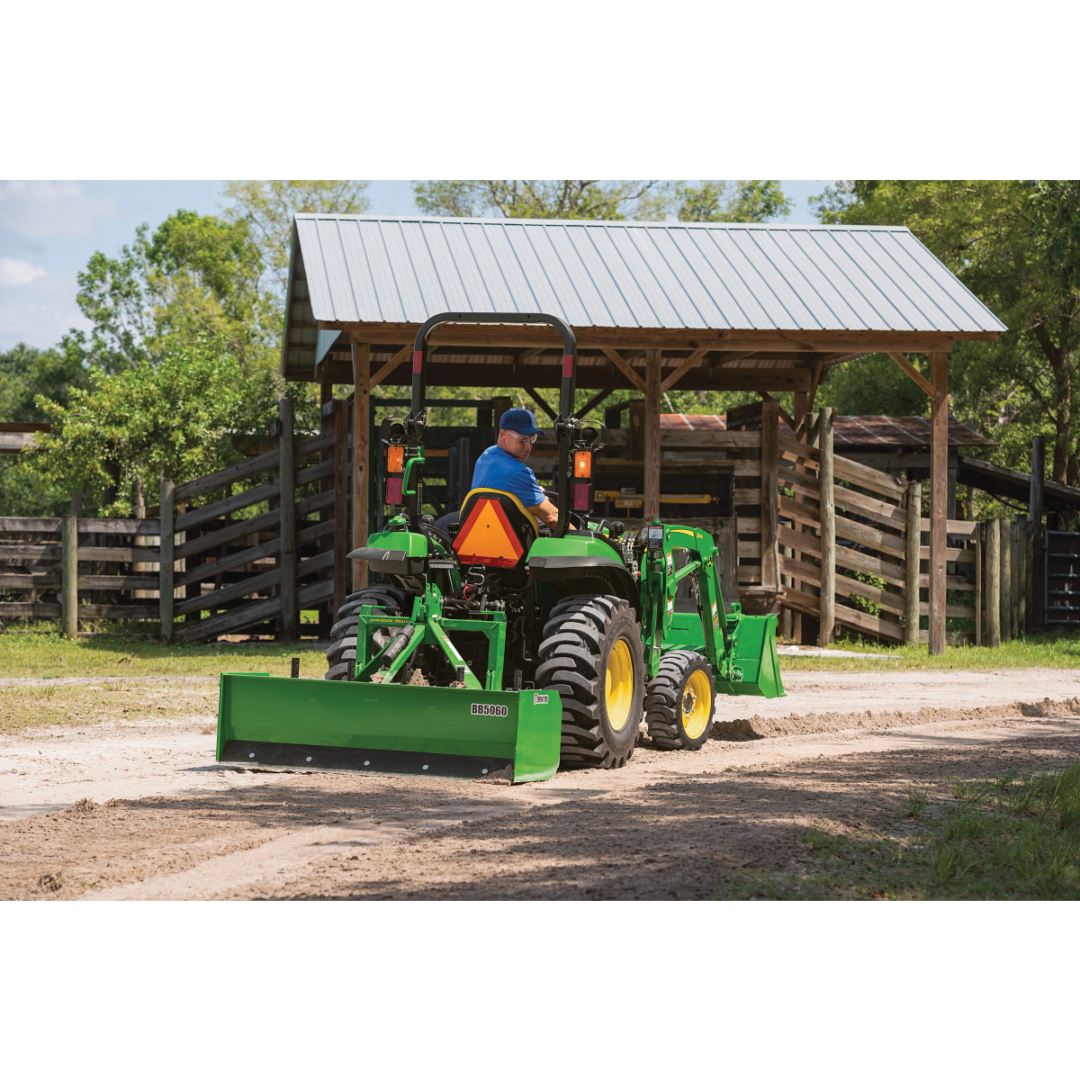 Person driving a green tractor with a front loader on a gravel path near a wooden shed and fenced paddock, trees in the background, on a sunny day