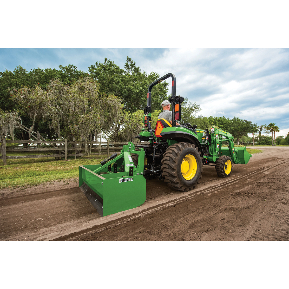 Green compact tractor with large rear tires and a rear grading box drives along a dirt path in a rural setting, bordered by grass, trees, and a wooden fence under a cloudy sky