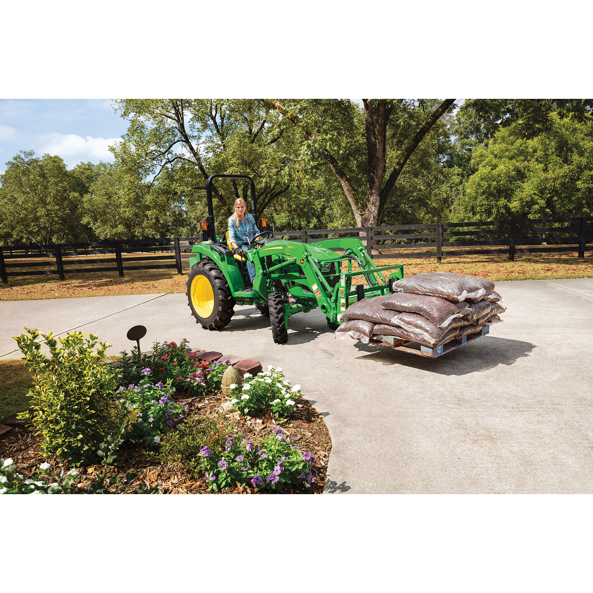 A green tractor with a front loader carries a pallet of bagged mulch across a paved driveway, with an operator seated at the controls, landscaped flowers in the foreground, and trees and fencing in the background