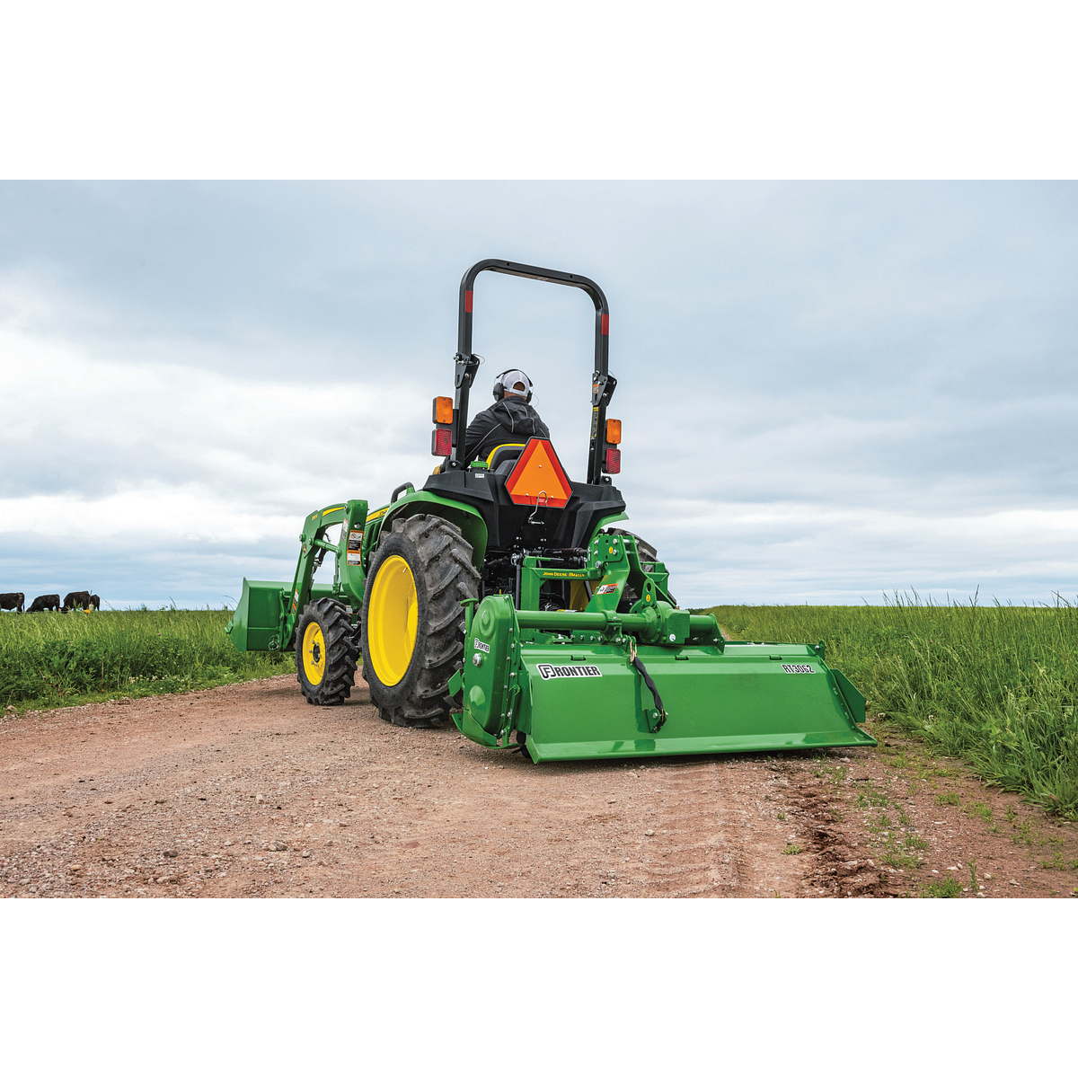 Rear view of a compact tractor driving on a dirt path with a rotary tiller attached, working along a grassy field under a cloudy sky