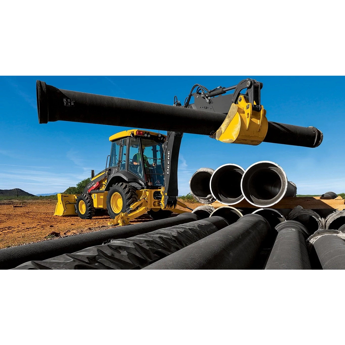 310 G-Tier Backhoe Loader carrying a piece of pipe to a pile for pipeline construction in large open field
