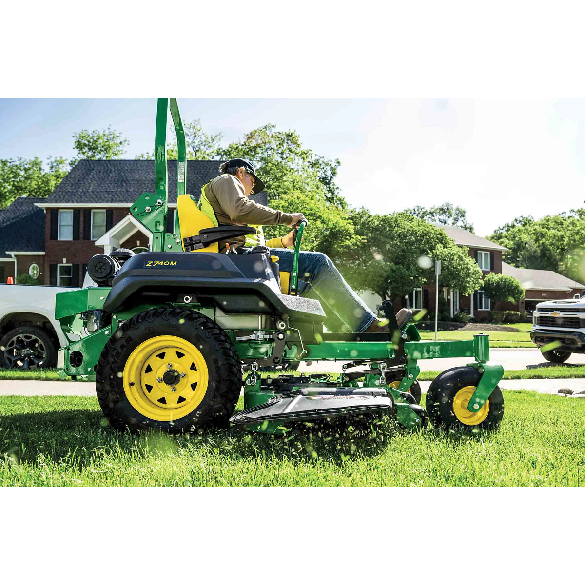 A landscaper operating a green zero-turn mower in a residential area with blades of cut grass coming towards the camera.