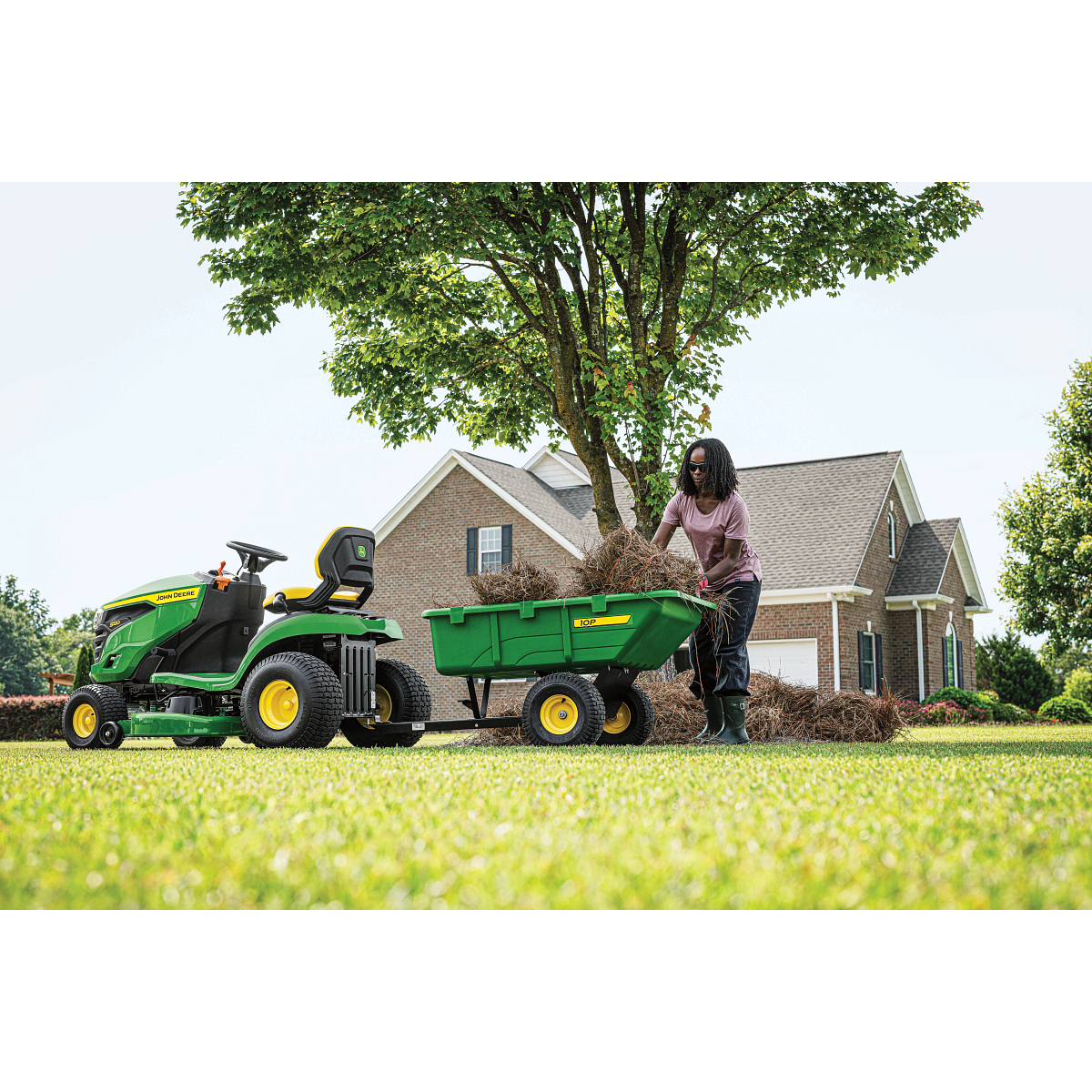 Side left view of a homeowner loading brush into the green 10P trailer attached to an S120 Lawn Tractor