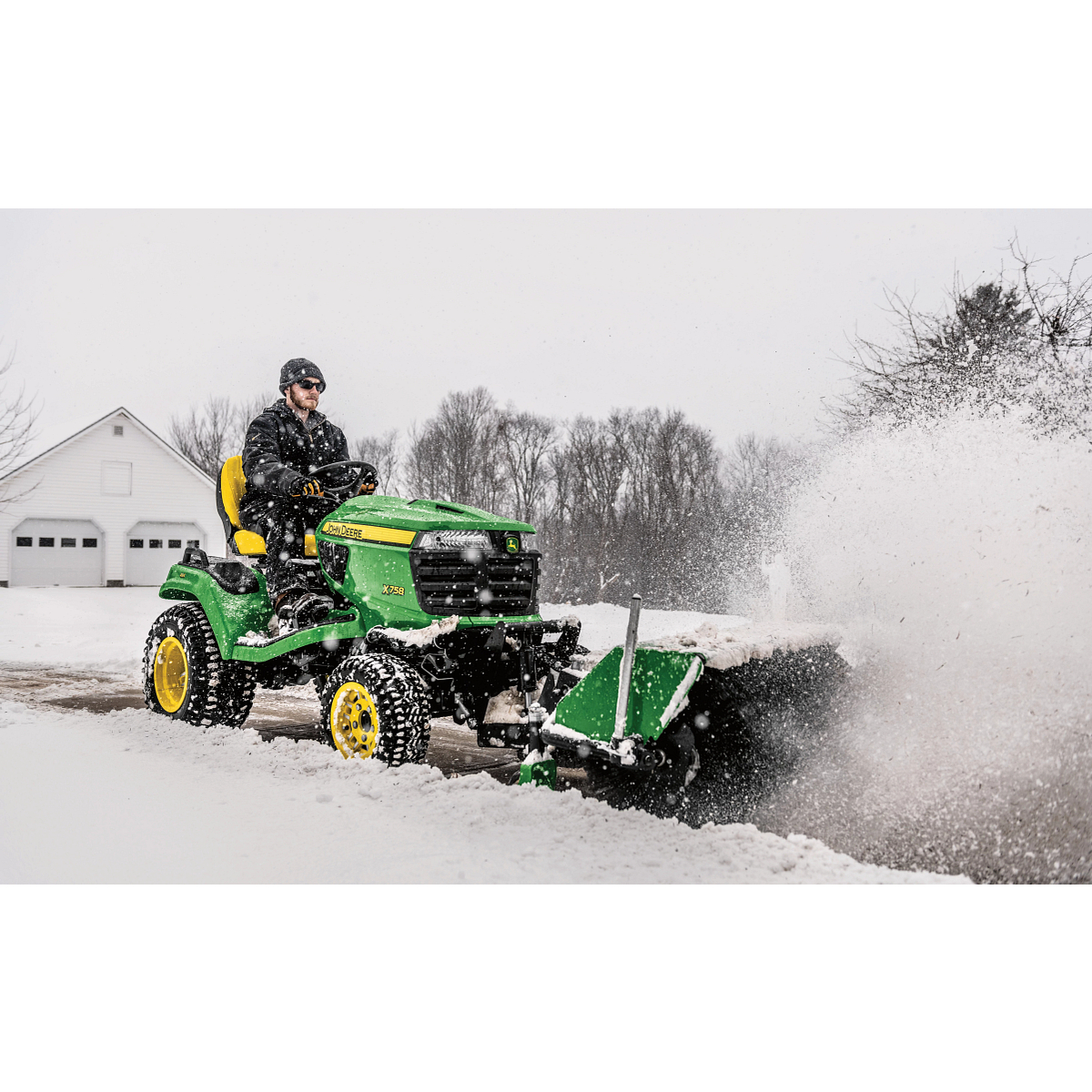 Front right view of an operator on an X758 lawn tractor blowing snow from a driveway with front-attached 52-in. rotary broom