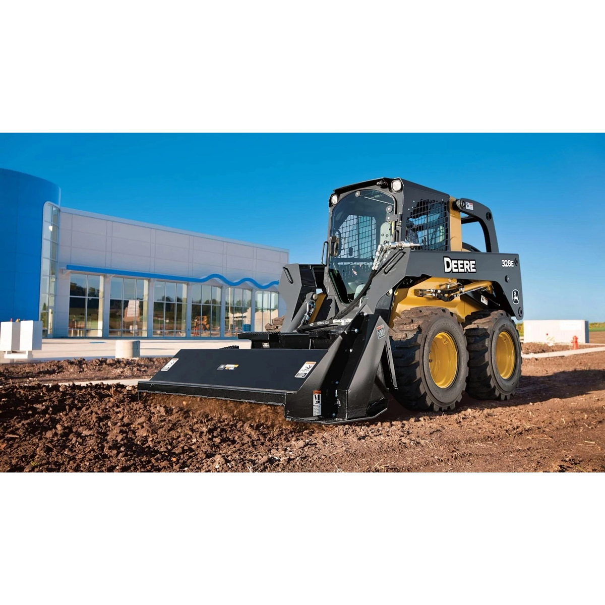 Front left view of 328E Skid Steer Loader with RT Series Rotary Tiller tilling fresh dirt in front of a large building