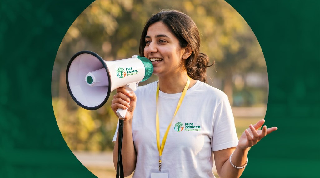 Smiling young woman wearing a white Pure Zameen Foundation t-shirt and yellow lanyard speaking into a megaphone outdoors with blurred trees in the background.