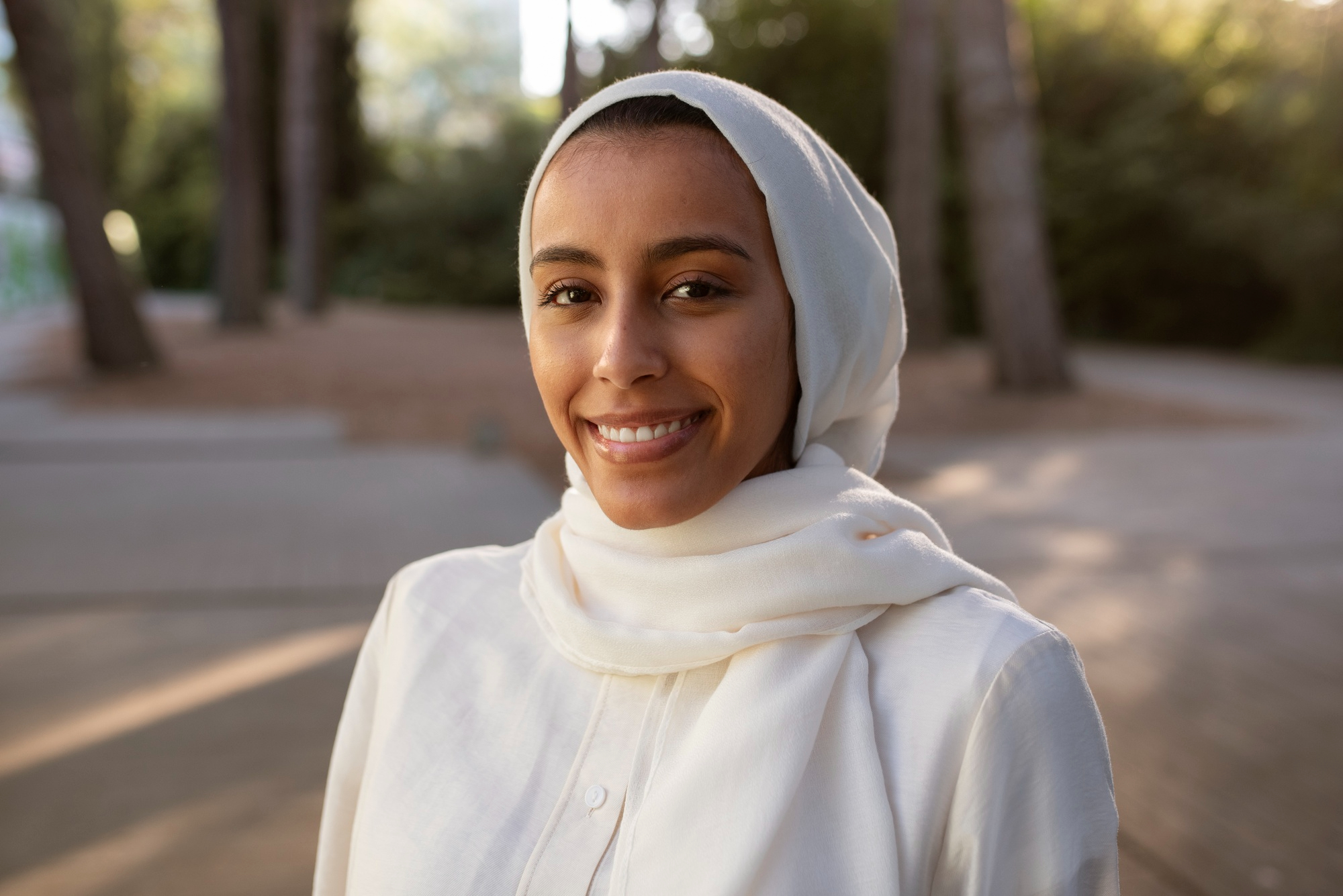 Smiling woman wearing a white hijab and white shirt outdoors with blurred trees and walkways in the background.