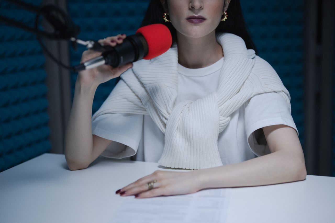 A woman wearing a white shirt and a white sweater draped over her shoulders holds a microphone with a red foam cover in a soundproof room, sitting at a white table with a paper in front of her.