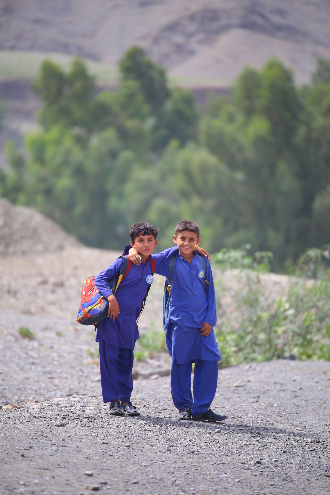 Two boys in matching blue traditional clothing standing on a rocky dirt path with one boy's arm around the other's shoulder, both carrying backpacks, with trees and hills blurred in the background.