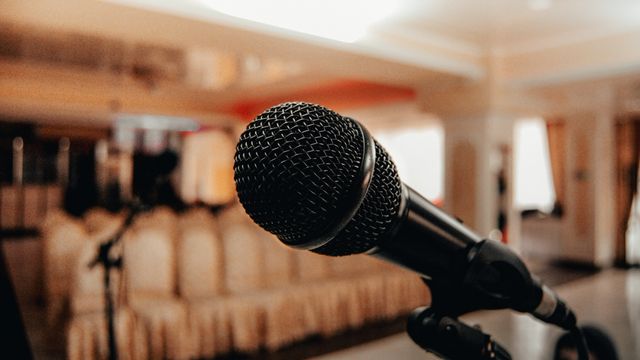 Close-up of a black microphone on a stand in an indoor setting with blurred brown chairs and warm lighting in the background.