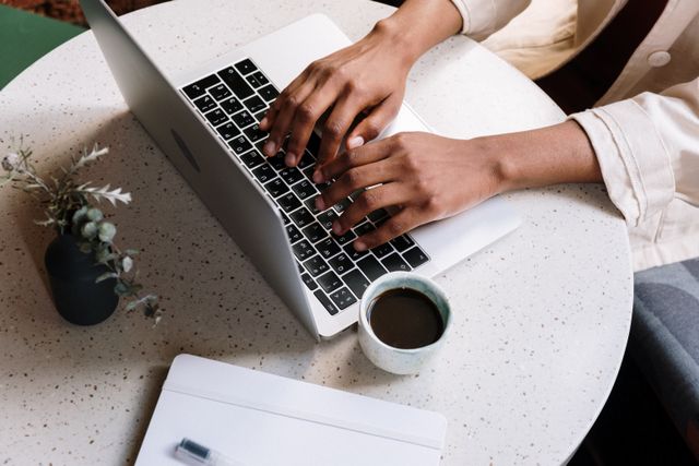 Person typing on a laptop keyboard at a round white speckled table with a cup of coffee, a closed notebook, and a vase with dried flowers.