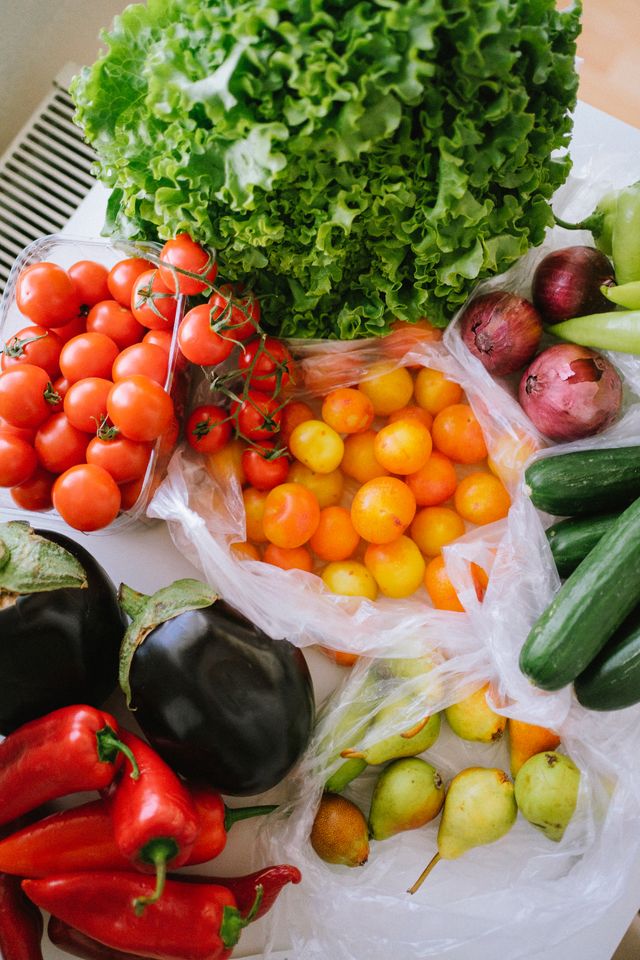 A variety of fresh vegetables and fruits on a table, including leafy green lettuce, red cherry tomatoes, yellow and orange cherry tomatoes in a plastic bag, two eggplants, red chili peppers, green cucumbers, red onions, and green pears in a plastic bag.