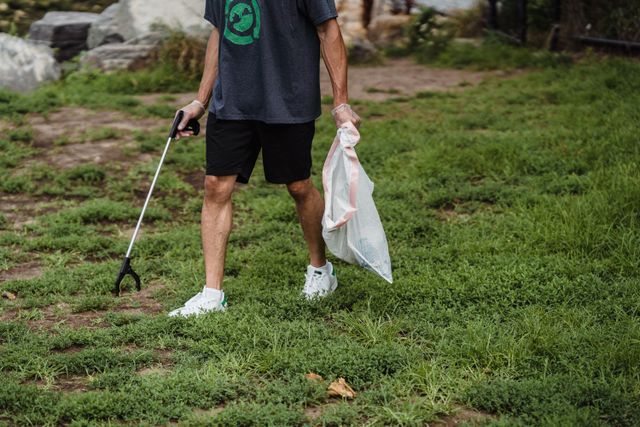 Person wearing a gray shirt and black shorts picking up trash with a litter picker in one hand and holding a plastic bag with collected trash in the other, standing on a grassy area.