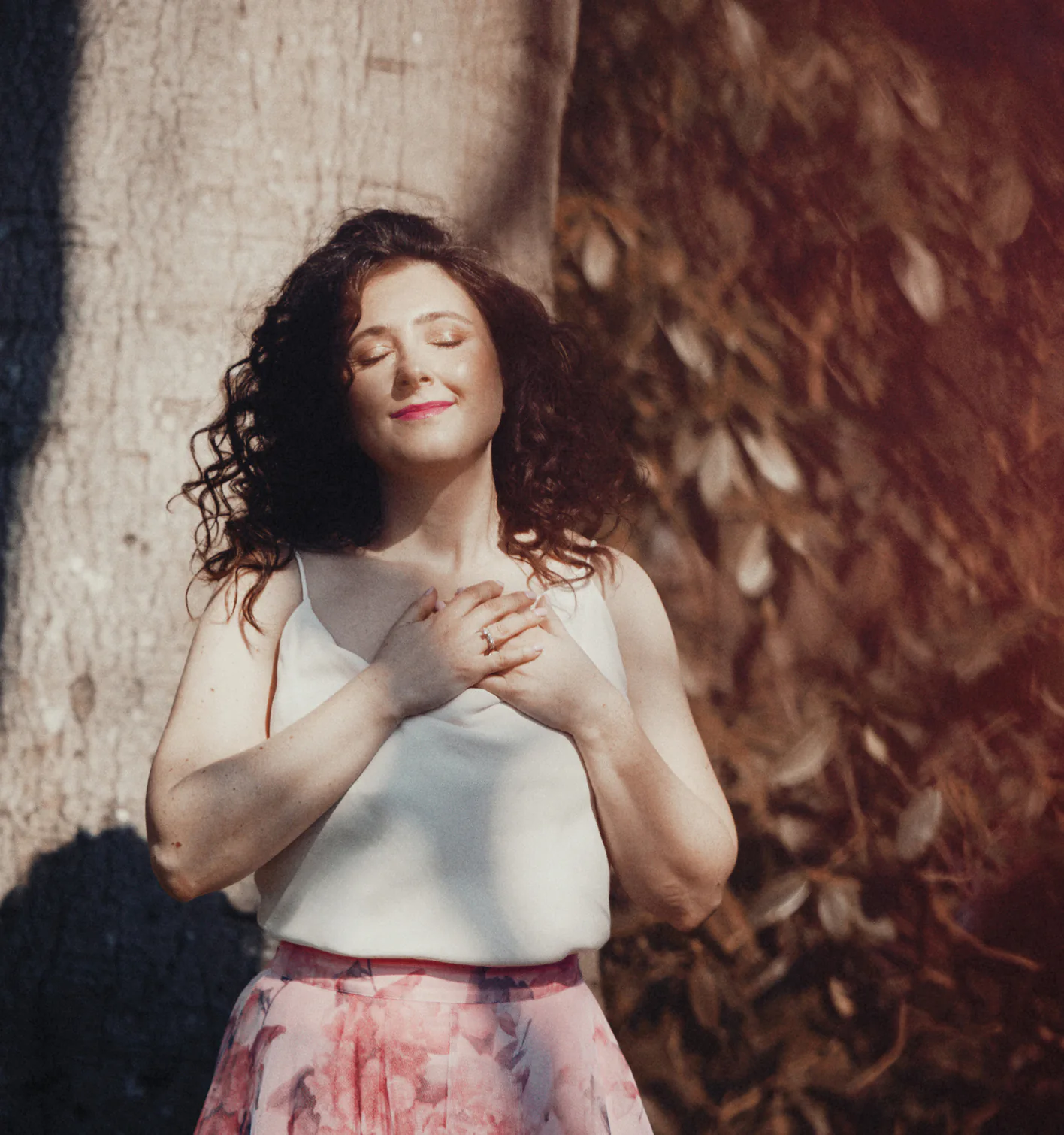 Woman in a white top and pink floral skirt standing outdoors with eyes closed and hands on chest.