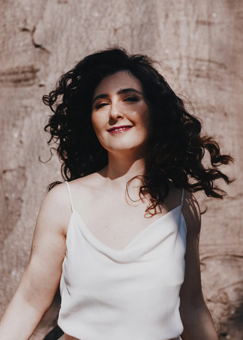 Smiling woman with curly dark hair wearing a white sleeveless top against a textured wall.