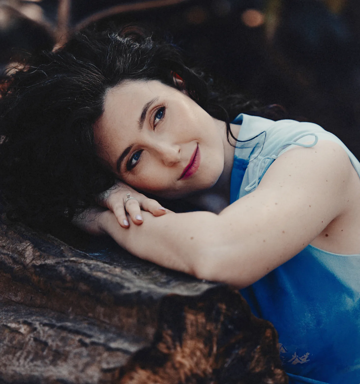 Woman with dark curly hair resting her head on folded arms on a log, wearing blue sleeveless top.