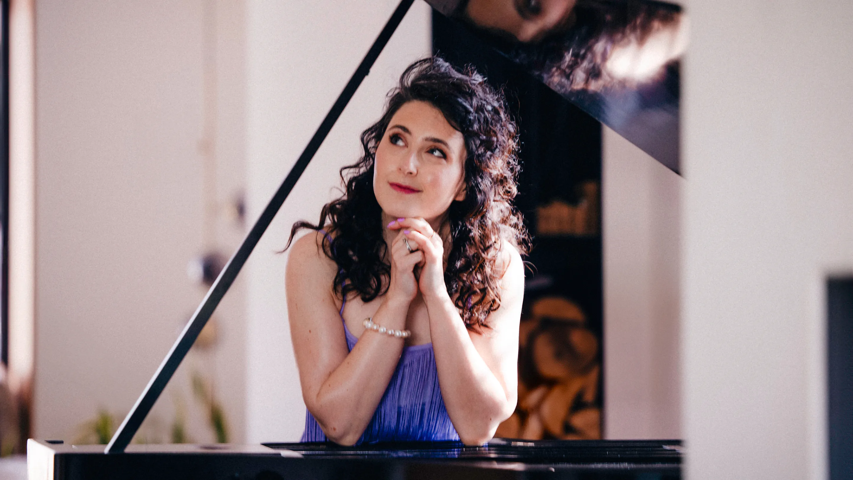 Woman with curly hair in a purple dress leaning on a grand piano, looking upward thoughtfully.