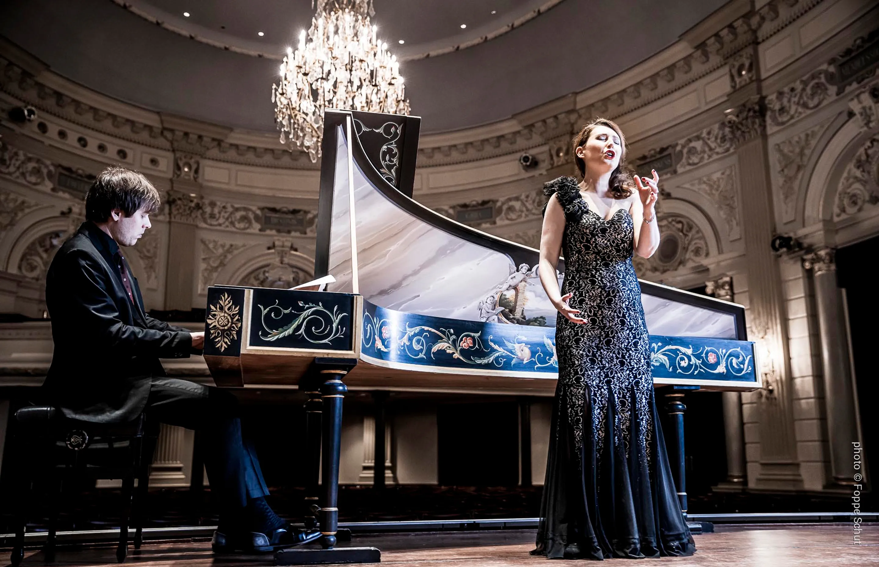 Woman in elegant gown singing next to a man playing a decorated harpsichord under a chandelier.
