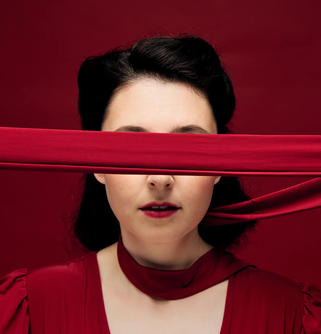 Woman in red dress with dark hair, blindfolded by a red cloth across her eyes against a red background.
