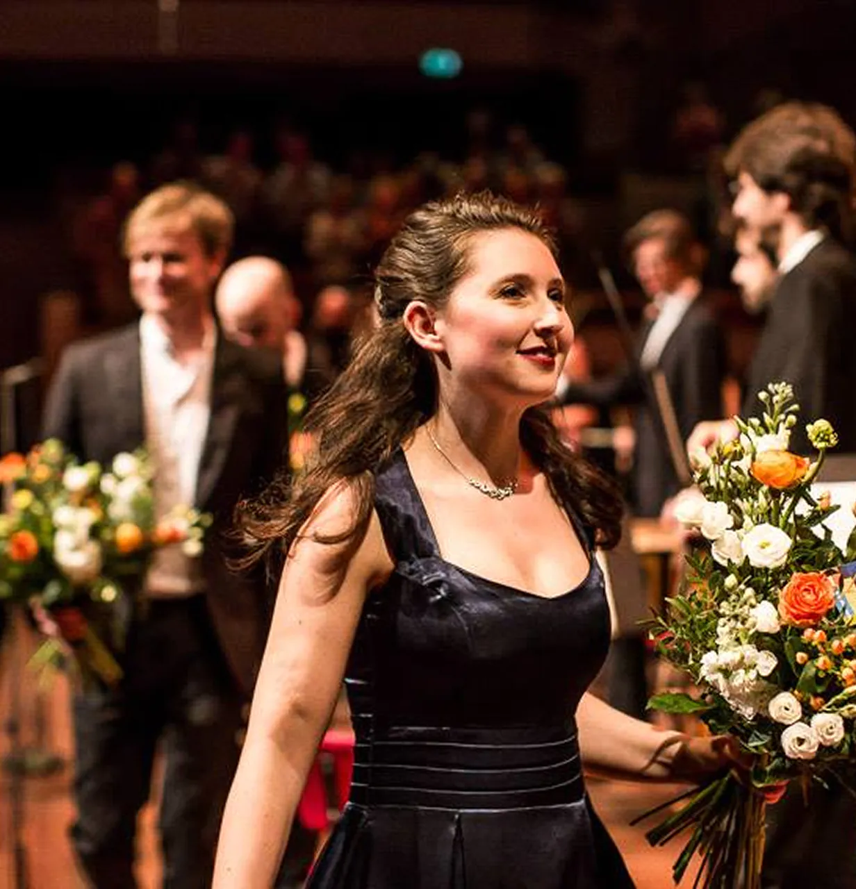 Woman in a black dress holding a bouquet of flowers, with people and bouquets blurred in the background.