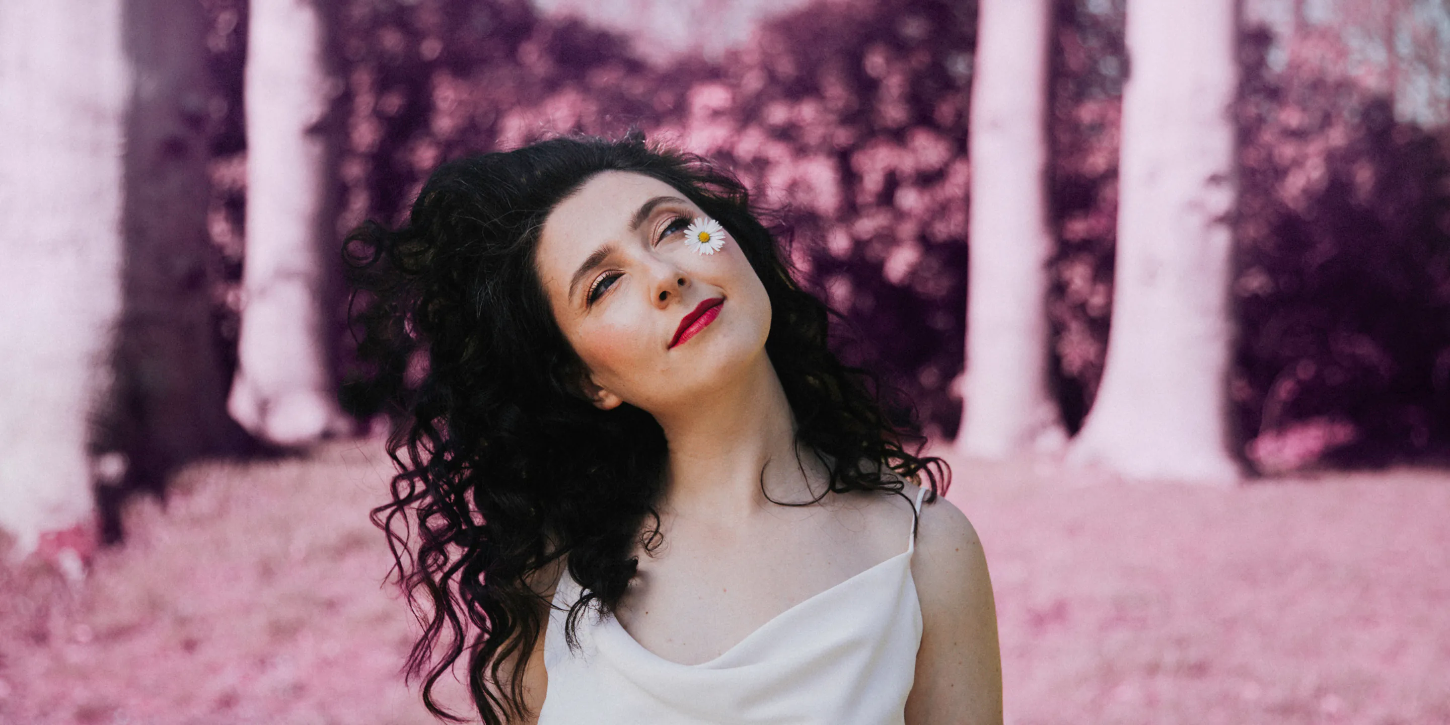 Woman with curly black hair and red lipstick with a daisy near her eye in a pink-toned forest.