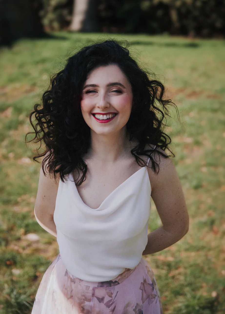 Smiling woman with curly dark hair wearing a white top and floral skirt outdoors on grass.