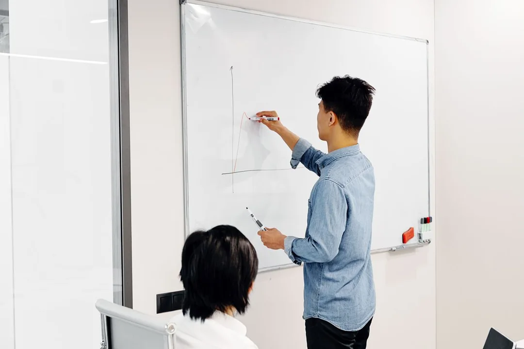 A colleague sketching a chart on a whiteboard while another looks on in a bright office