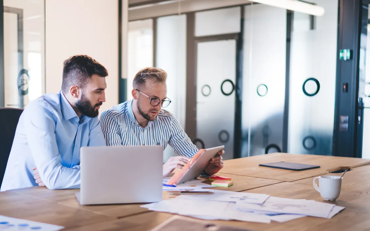 Two colleagues working through findings on a laptop and tablet at a meeting table