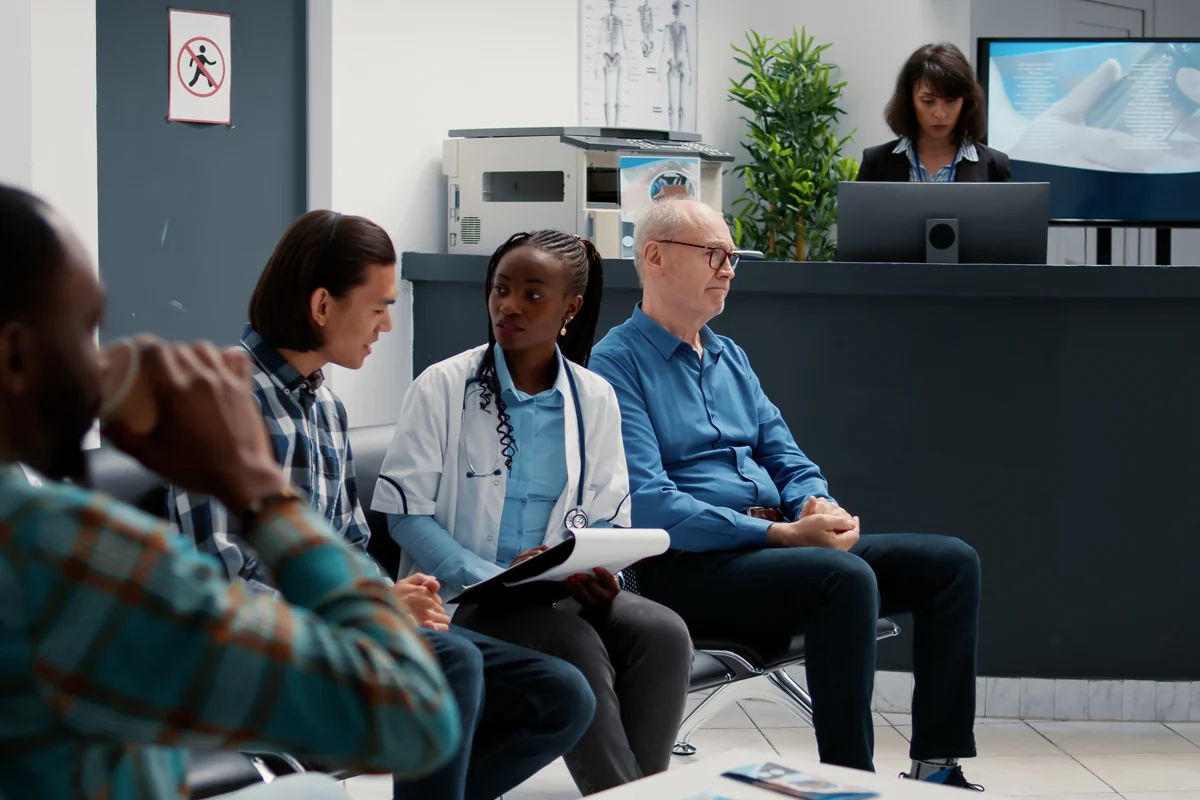 Patients waiting in a clinic reception area while a staff member works at the front desk behind them