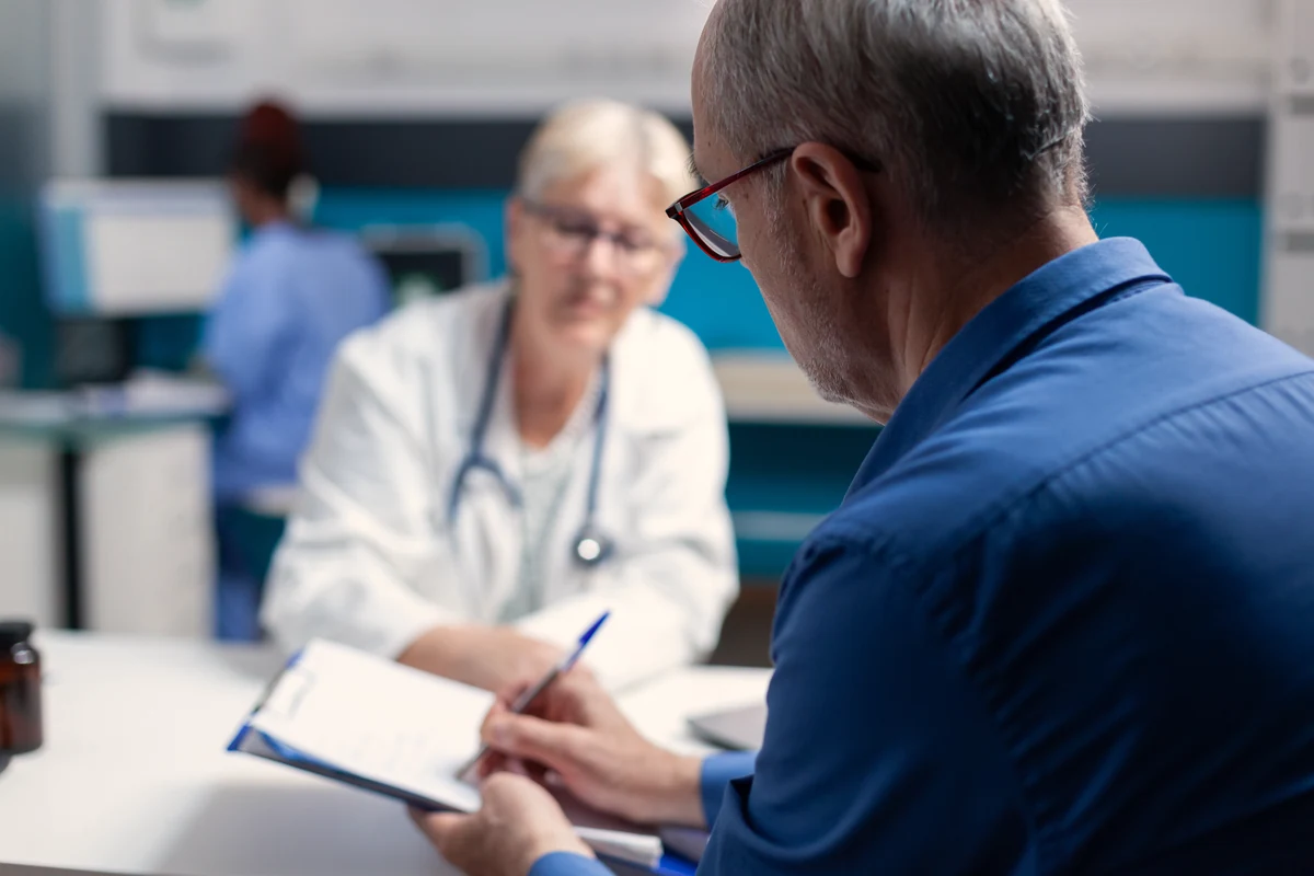 A patient completing a form on a clipboard at a medical practice while staff work behind him